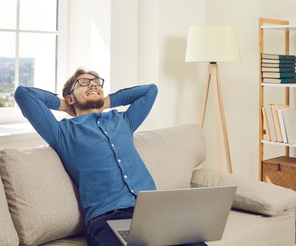 Happy calm young man sit on sofa relax after work finish on laptop. Smiling Caucasian male rest on couch in living room take break from computer job breathe fresh air. Freelance, relaxation concept.